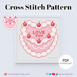Può includere: Uno schema di punto croce di una torta rosa a forma di cuore con glassa bianca e ciliegie rosse. La torta dice "Love You" al centro.
