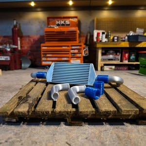Könnte beinhalten: Ein miniaturisierter, silberfarbener Ladeluftkühler für Kraftfahrzeuge mit blauen Silikonkupplungen und zwei silberfarbenen Metallrohrbögen. Der Ladeluftkühler verfügt über ein horizontales Lamellendesign und wird mit separaten blauen Verbindungsmuffen gezeigt.