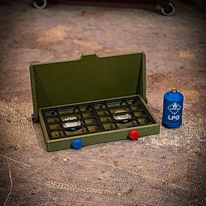 Puede incluir: Una estufa de camping en miniatura de color verde oliva con dos quemadores, perillas rojas y azules y una rejilla negra. Un tanque de GLP azul está al lado de la estufa. El fondo incluye un cofre de herramientas naranja.