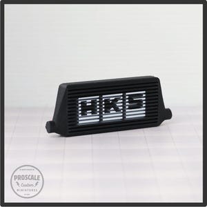May include: A black miniature intercooler featuring the white letters "HKS". The model has a ribbed design and is a custom miniature. The product is a Proscale Custom Miniature.