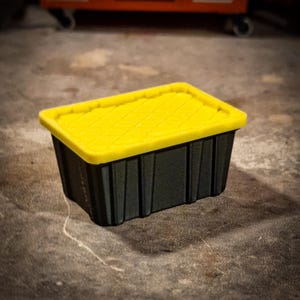 May include: A black storage container with a bright yellow lid. The lid has a textured surface, and the container has vertical ridges on the sides. Ideal for organising and storing items.