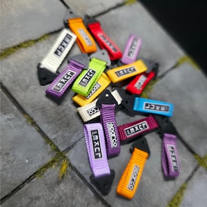 Puede incluir: Una colección de correas de remolque de coche de tela de colores y marcas, con logotipos como Sparco y Maxpider. Las correas están hechas de nylon tejido y tienen un lazo en un extremo y un gancho de metal en el otro.