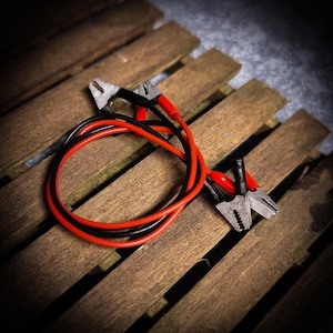 Puede incluir: Cables de puente rojos y negros con pinzas plateadas, colocados sobre una superficie de madera. Estos cables se utilizan para arrancar un vehículo con la batería descargada.