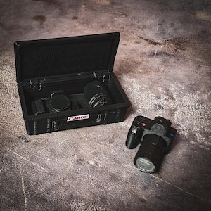 May include: A black miniature camera and lens set, with a matching case. The case is open, revealing the camera and lens inside. The word "Canon" is printed on the case. The camera is next to the case.