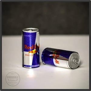 Könnte beinhalten: Zwei Miniatur-Red Bull-Energy-Drink-Dosen. Die Dosen sind silberfarben mit blauen und roten Grafiken. Der Text "Red Bull" und "Energy Drink" ist auf den Dosen sichtbar.
