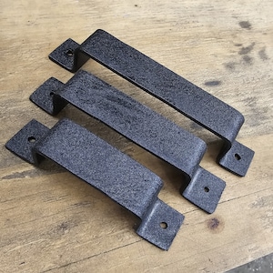 Może przedstawiać: Trzy czarne metalowe uchwyty do drzwi z fakturowanym wykończeniem. Uchwyty są prostokątne ze zaokrąglonymi krawędziami i mają jeden otwór na każdym końcu.