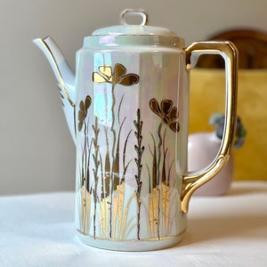 May include: A white ceramic teapot with a gold handle and floral design. The teapot has a pearlescent glaze and features gold accents.