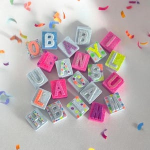 May include: A collection of colorful, glittery letter tiles spelling out "BABY CAM IS LULA". The tiles are made of a light blue material and have a variety of colors and glitter.