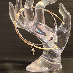 May include: A white and pink wire wrapped bracelet with a silver charm in the shape of a cone. The bracelet is displayed on a clear plastic hand.