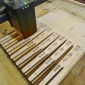 May include: A laser cutter engraving wooden fans. The fans are light brown with decorative cutouts and the text "KEO STUDIO S&G 091224". The laser cutter is black with green, blue, and red lights.