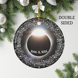 May include: A double-sided, round Christmas ornament featuring a solar eclipse design. The ornament has a black background with a bright sun and a ring of white speckles. The text "APRIL 8, 2024" is displayed in white.