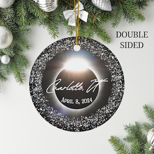 May include: A black and white round ornament with a silver glitter border. The ornament features a solar eclipse with the text "Charlotte, TX" and "April 8, 2024".