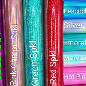 May include: A collection of holographic and sparkly vinyl rolls in various colors including purple, pink, green, red, blue, silver, emerald, white opal, and hot pink. Each roll is labeled with text such as Purple Spkl, Pink Chmpn Spkl, Green Spkl, Red Spkl, Peacock Holo, Silver Holo, Emerald Hol, White Opal Holo, and Hot Pink Holo.