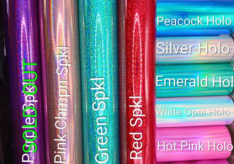 May include: A collection of holographic and sparkly vinyl rolls in various colors including purple, pink, green, red, blue, silver, emerald, white opal, and hot pink. Each roll is labeled with text such as Purple Spkl, Pink Chmpn Spkl, Green Spkl, Red Spkl, Peacock Holo, Silver Holo, Emerald Hol, White Opal Holo, and Hot Pink Holo.
