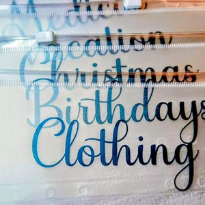 May include: A stack of clear, translucent plastic zipper pouches with white plastic sliders. Each pouch features black cursive text printed on the front, including the words Medication, Vacation, Christmas, Birthdays, and Clothing, arranged in a vertical layout.