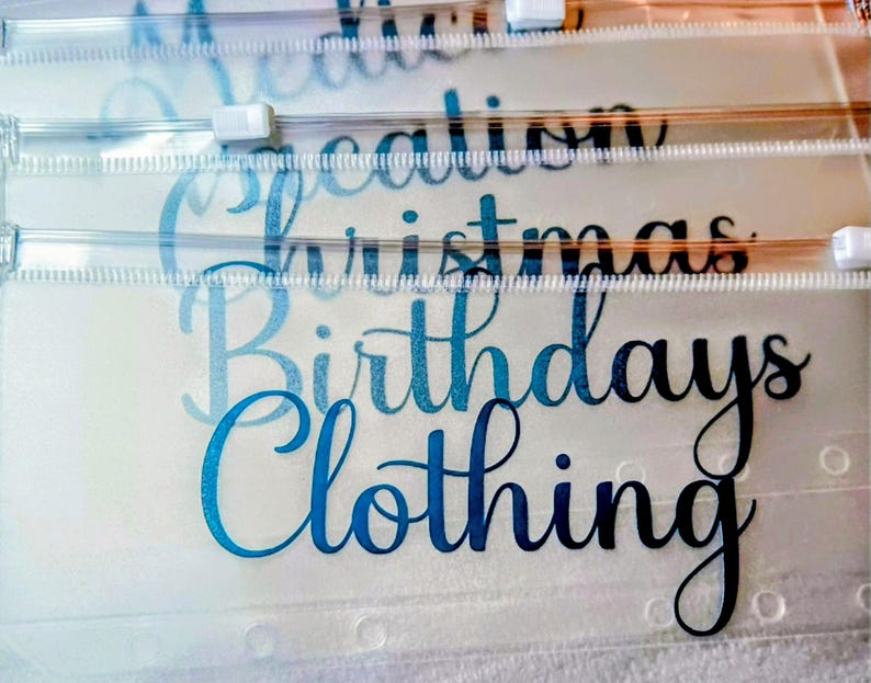 May include: A stack of clear, translucent plastic zipper pouches with white plastic sliders. Each pouch features black cursive text printed on the front, including the words Medication, Vacation, Christmas, Birthdays, and Clothing, arranged in a vertical layout.