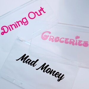May include: Three clear plastic zipper pouches for binders. One pouch features the hot pink text Dining Out, another has light pink text reading Groceries, and the third displays black cursive text that reads Mad Money. Each pouch has holes.