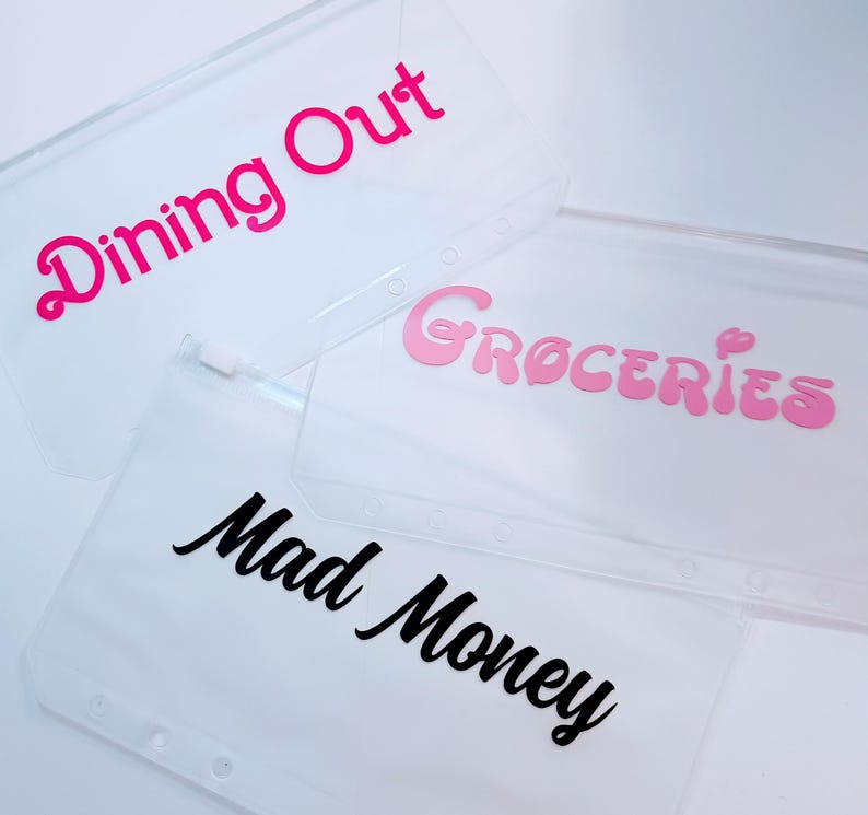 May include: Three clear plastic zipper pouches for binders. One pouch features the hot pink text Dining Out, another has light pink text reading Groceries, and the third displays black cursive text that reads Mad Money. Each pouch has holes.