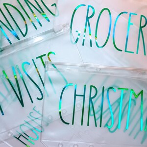 May include: A collection of clear plastic zipper pouches featuring iridescent green text. The pouches are labeled with words including SPENDING, GROCERIES, VET VISITS, and CHRISTMAS. Each pouch has a white zipper closure and punched holes along the side.