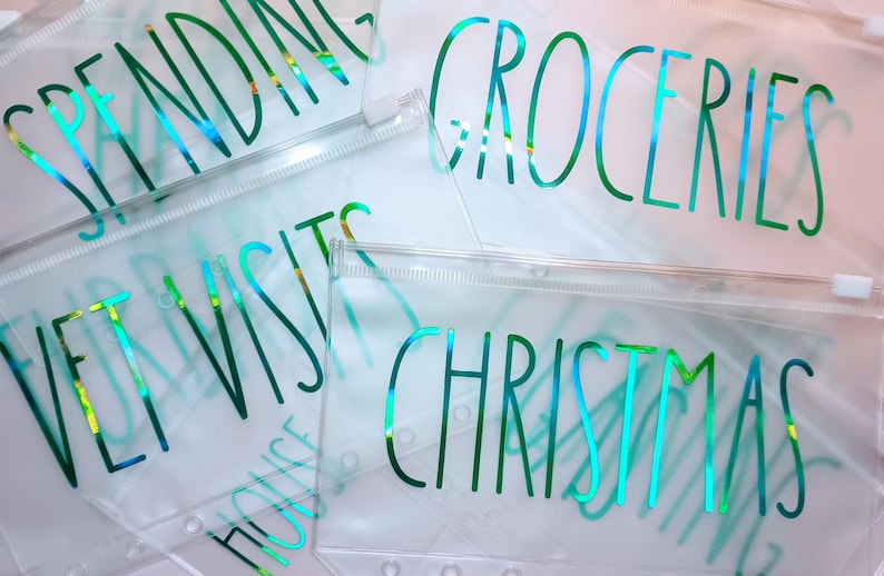 May include: A collection of clear plastic zipper pouches featuring iridescent green text. The pouches are labeled with words including SPENDING, GROCERIES, VET VISITS, and CHRISTMAS. Each pouch has a white zipper closure and punched holes along the side.