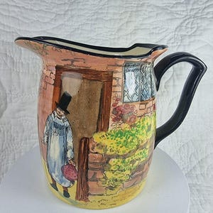 May include: A ceramic jug featuring a hand-painted scene of a person in a top hat standing by a doorway. The jug has a glossy finish, a black handle, and a black rim. The illustration includes a stone wall, a window, and foliage.