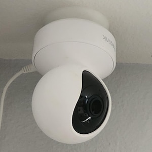 Könnte beinhalten: Eine weiße, kuppelförmige Überwachungskamera, die an einer Decke montiert ist. Die Kamera hat ein schwarzes Objektiv und ein abgerundetes, kugelförmiges Design. Ein weißes Kabel erstreckt sich von der Kamera.