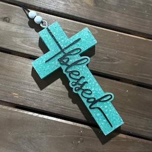 May include: A turquoise cross-shaped ornament with the word "Blessed" in black script. The ornament has a textured surface and is suspended by a black cord with white beads.