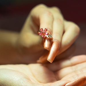 May include: A close-up shot of a flower-shaped ring with pink petals and small clear stones. The ring is held between two hands with long, polished nails. The background is a soft, pink fabric.