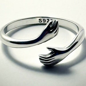May include: Silver ring with two hands clasped together, forming a circle. The ring is marked with the number "S925" on the inside.