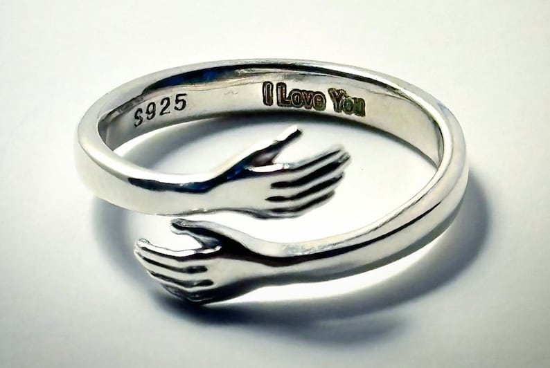 K&ouml;nnte beinhalten: Silberring mit zwei ineinander verschlungenen H&auml;nden, die eine Umarmung bilden. Der Ring ist mit "I Love You" und "S925" graviert.
