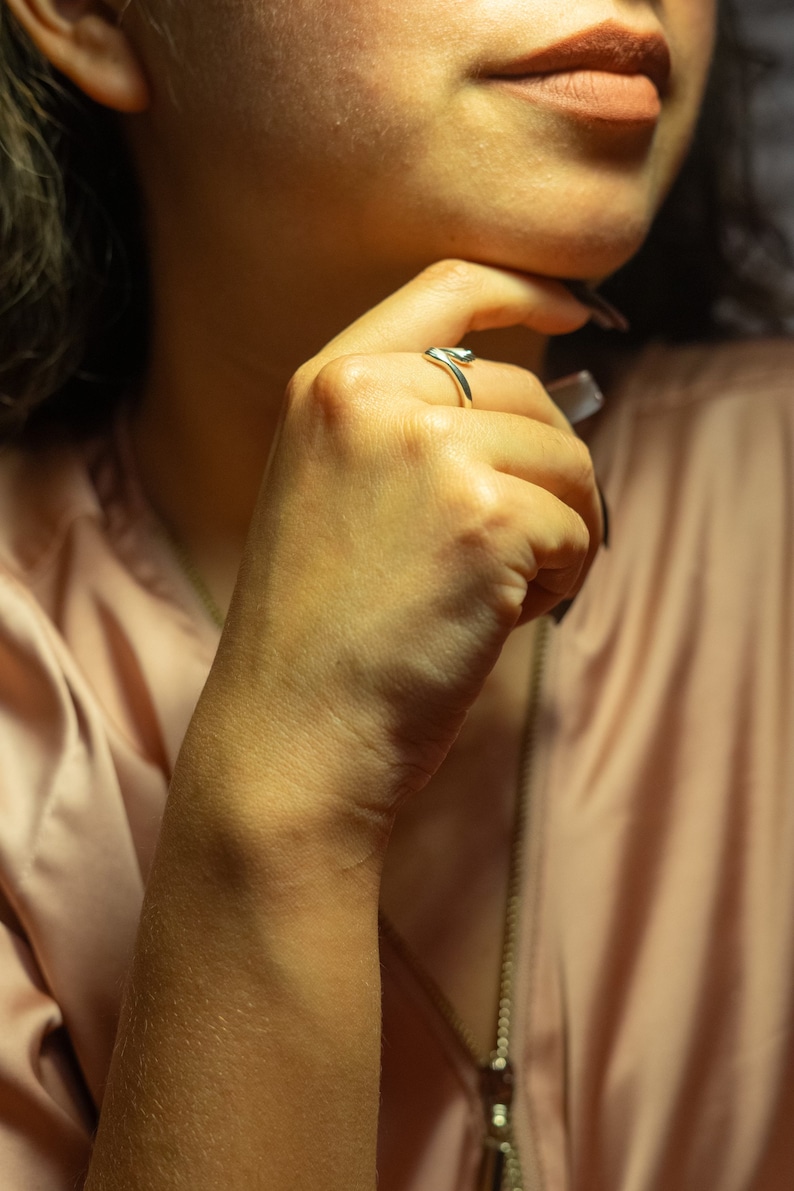 K&ouml;nnte beinhalten: Nahaufnahme einer Hand, die einen silbernen Ring tr&auml;gt. Der Ring ist ein schlichtes Band mit einem geschwungenen Design. Die Hand der Person ruht auf ihrem Kinn, und sie tr&auml;gt ein hellbraunes Hemd.