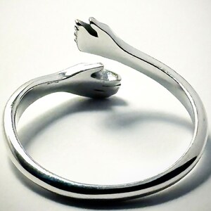 K&ouml;nnte beinhalten: Ein silberner Ring mit zwei ineinander verschlungenen H&auml;nden, die einen Kreis bilden. Der Ring ist aus Metall und hat eine glatte, polierte Oberfl&auml;che.