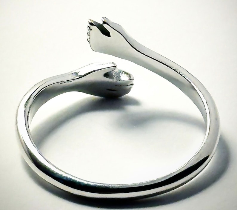 K&ouml;nnte beinhalten: Ein silberner Ring mit zwei ineinander verschlungenen H&auml;nden, die einen Kreis bilden. Der Ring ist aus Metall und hat eine glatte, polierte Oberfl&auml;che.