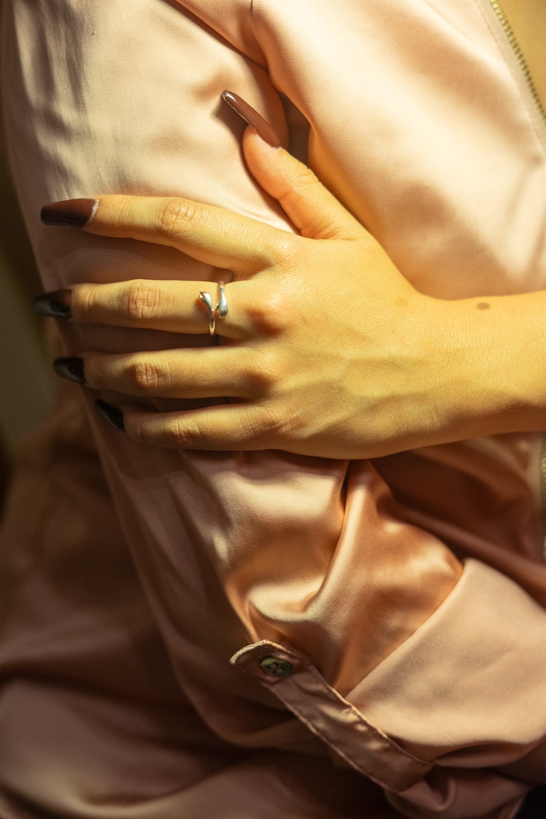K&ouml;nnte beinhalten: Nahaufnahme einer Hand, die einen silbernen Ring mit einem einzigartigen Design tr&auml;gt. Die Hand ruht auf einem weichen, pfirsichfarbenen Stoff.