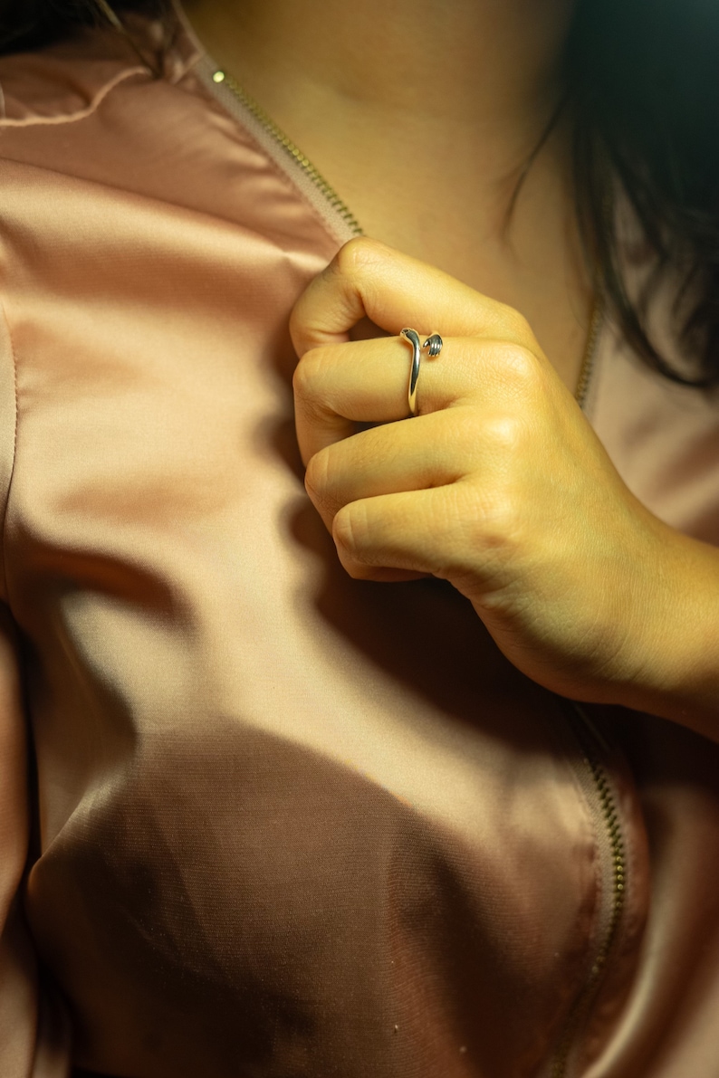 K&ouml;nnte beinhalten: Nahaufnahme einer Hand, die einen silbernen Ring mit einem einzigartigen Design tr&auml;gt. Der Ring befindet sich am Mittelfinger und besteht aus einem d&uuml;nnen, gebogenen Band mit einem kleinen, runden Detail oben.