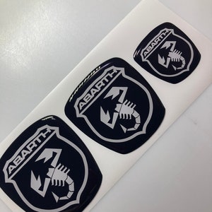 Pode incluir: Três emblemas de carro pretos e prateados com a palavra "ABARTH" e um logotipo de escorpião.