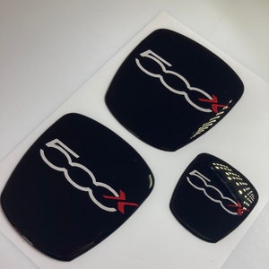 Può includere: Tre emblemi di auto neri e bianchi con una croce rossa. Gli emblemi hanno la forma di rettangoli arrotondati e riportano il testo "500" in bianco con un design a catena bianca.