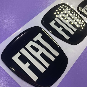 Può includere: Tre emblemi di auto FIAT neri e bianchi su un foglio di supporto bianco. Gli emblemi sono lucidi e hanno un effetto 3D.