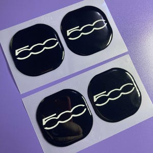 Könnte beinhalten: Vier schwarze, glänzende Autoembleme mit der weißen Zahl "500". Die Embleme sind oval geformt und befinden sich auf einer weißen Unterlage. Der Hintergrund ist helllila.
