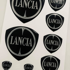 Può includere: Un set di sette emblemi per auto neri e argentati con la scritta "LANCIA" in lettere bianche.