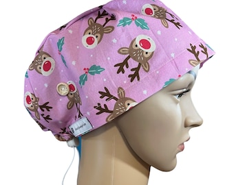 May include: A pink surgical scrub cap featuring a repeating pattern of brown reindeer with red noses and green holly leaves. The cap has a drawstring and a small tag with text. The cap is designed to be worn in a medical setting.
