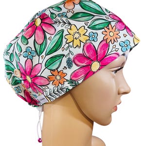 May include: A white scrub cap with a colorful floral print featuring pink, yellow, orange, and blue flowers. The cap has a drawstring and a button on the side.