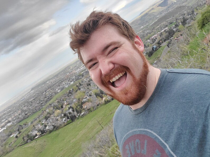 May include: A man with a red beard is laughing with his mouth open, showing his teeth. He is wearing a blue t-shirt with a red circle and the word "LOVE" in white letters. The background is a blurry view of a city.