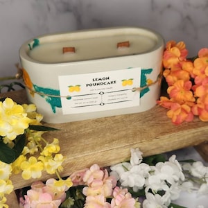 May include: A cream-coloured, oval candle with two wooden wicks, labelled "Lemon Poundcake." The container has teal and orange accents and twine. The candle is on a wooden surface, surrounded by yellow, pink, orange, and white flowers. The candle weighs 240g.