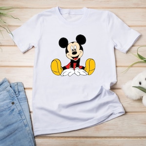 May include: White t-shirt with a cartoon image of Mickey Mouse sitting down and smiling. Mickey Mouse is wearing red shorts and yellow shoes.