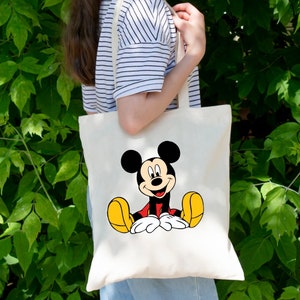 May include: A white canvas tote bag with a cartoon image of Mickey Mouse sitting down. Mickey Mouse is wearing red shorts and yellow shoes.