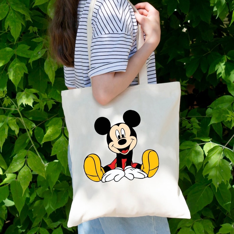 May include: A white canvas tote bag with a cartoon image of Mickey Mouse sitting down. Mickey Mouse is wearing red shorts and yellow shoes.