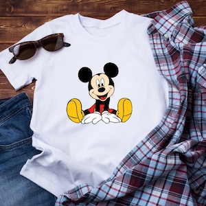 May include: White t-shirt with a cartoon image of Mickey Mouse sitting down. Mickey Mouse is wearing red shorts and yellow shoes.