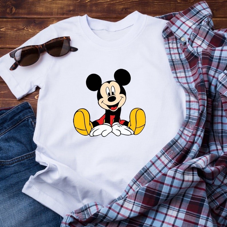 May include: White t-shirt with a cartoon image of Mickey Mouse sitting down. Mickey Mouse is wearing red shorts and yellow shoes.