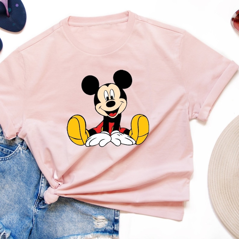May include: A pink t-shirt with a cartoon image of Mickey Mouse sitting down with his hands in his lap. Mickey Mouse is wearing red shorts and yellow shoes.